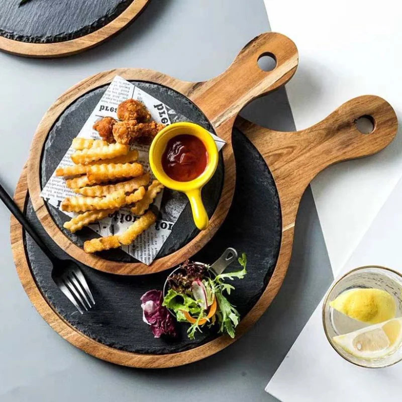 A styled overhead flat-lay of a round black slate serving board with a wooden handle, holding crinkle-cut fries, fried pieces, a small yellow cup of red sauce, and a small salad garnish on a gray surface.