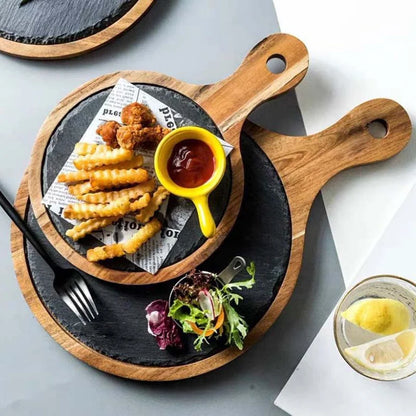 A styled overhead flat-lay of a round black slate serving board with a wooden handle, holding crinkle-cut fries, fried pieces, a small yellow cup of red sauce, and a small salad garnish on a gray surface.