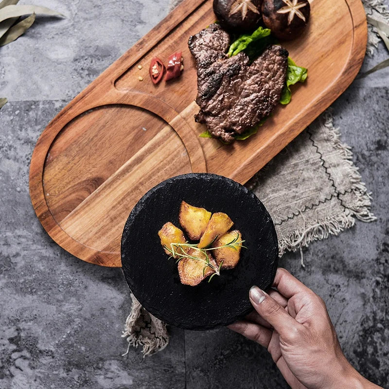 A hand holds a round black slate serving platter with roasted potatoes, while a wooden serving board with a steak and mushrooms sits on a gray stone surface.