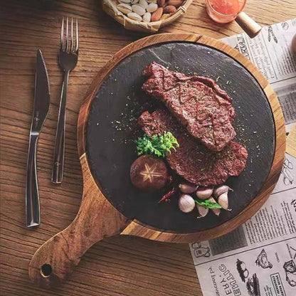A top-down view of a cooked steak served on a round black stone plate with a wooden handle, placed on a wooden table with a knife, fork, garlic cloves, a mushroom, herbs, and printed paper around it.