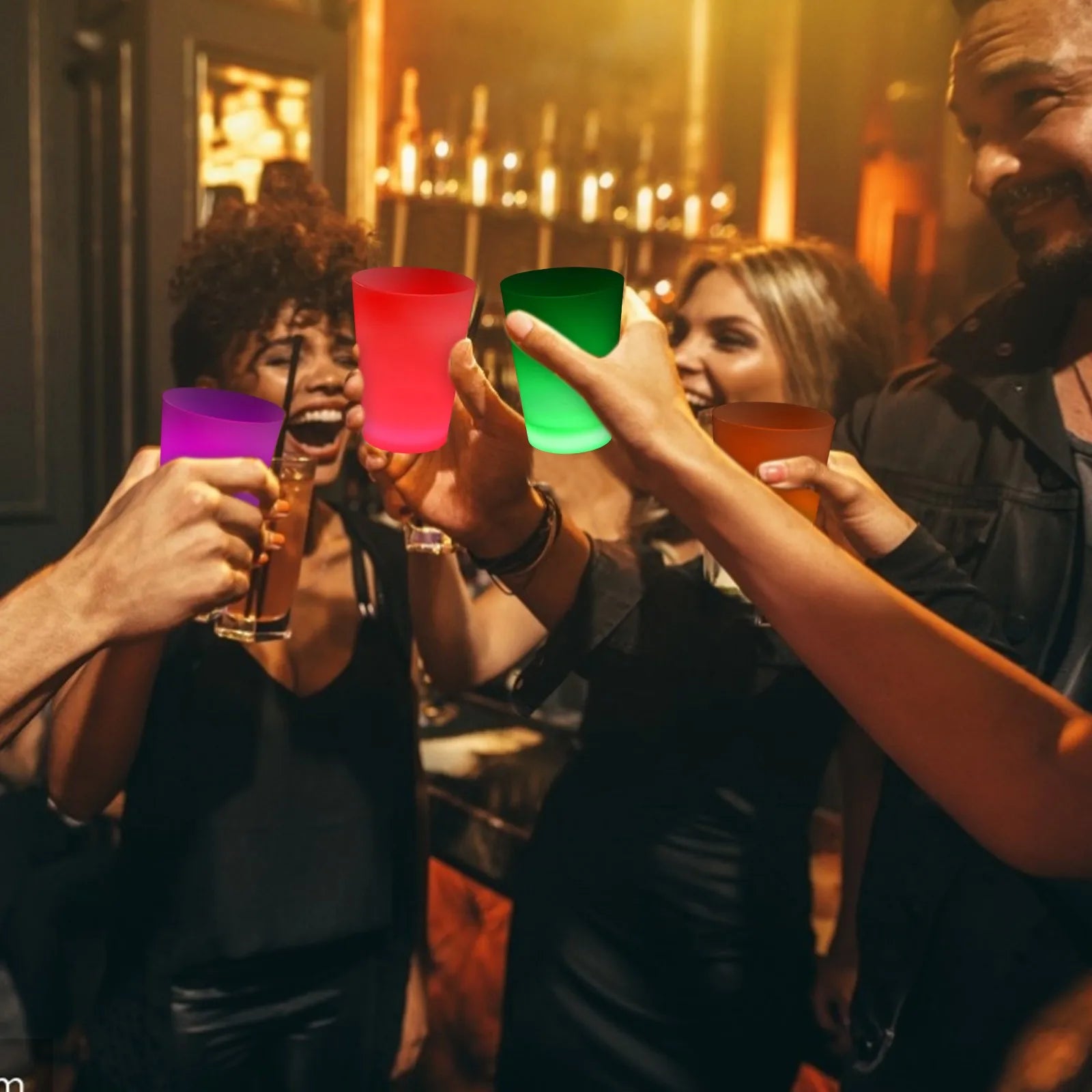 Three people in a dimly lit bar or restaurant setting raise colorful plastic cups in a toast. The cups are translucent and come in purple, red, green, and amber-brown shades, with warm ambient lighting reflecting off their surfaces.