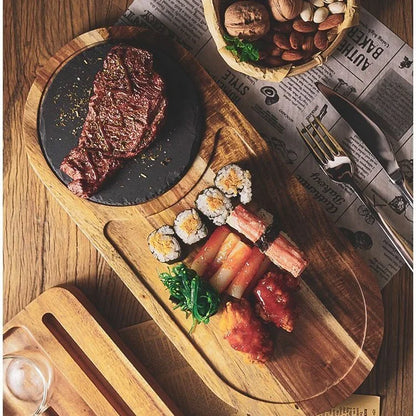 A styled overhead food presentation on a wooden board: a grilled steak on a black plate, sushi rolls, sliced pink fish or shrimp, red glazed pieces, greens, and a small basket of nuts on printed newspaper paper.