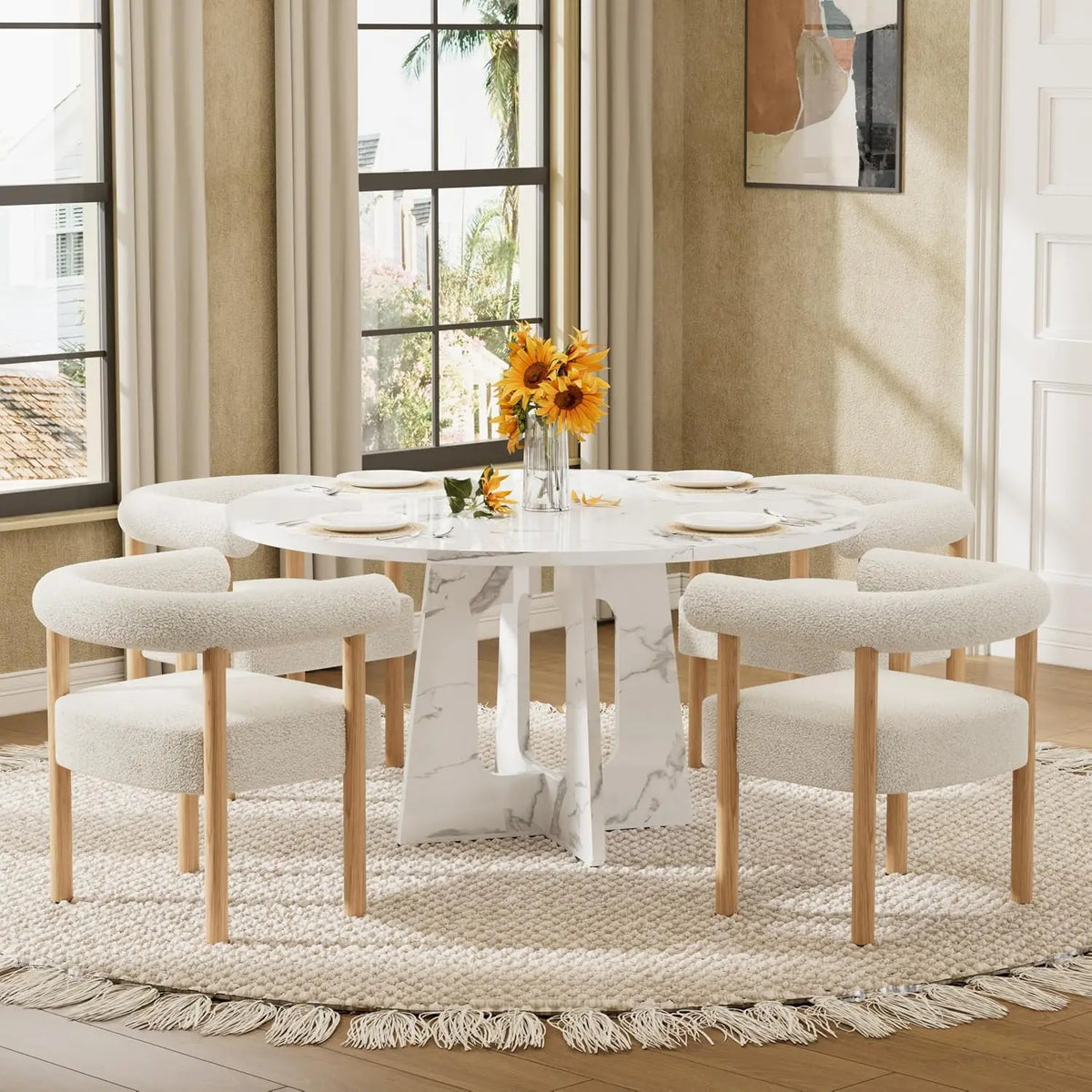 A round white marble dining table with a sculptural pedestal base, surrounded by six upholstered dining chairs with light wood legs, set on a woven fringe rug in a bright dining room.