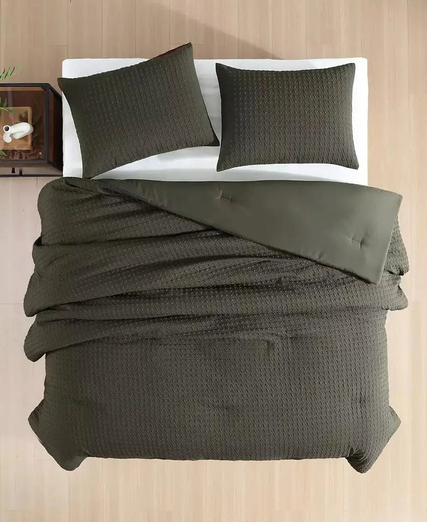 An overhead view of a bed dressed in an olive green textured waffle-weave bedding set, including a duvet cover and two matching pillowcases, set against a light wood-paneled background.