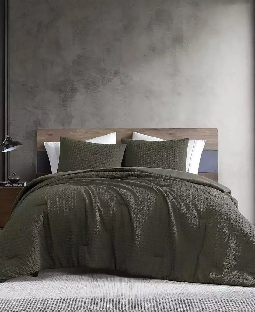An olive green textured bedding set, including a quilt and two matching pillow shams, styled on a bed with white pillows against a rustic gray concrete wall and wooden headboard.