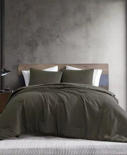 An olive green textured bedding set, including a quilt and two matching pillow shams, styled on a bed with white pillows against a rustic gray concrete wall and wooden headboard.