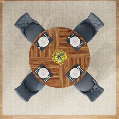Overhead view of a round wooden dining table with four gray tufted chairs, set on a beige rug with a wood floor visible around the edges. Each chair is placed opposite the table, with white plates, black napkins, silver cutlery, and a small yellow floral centerpiece on the table.