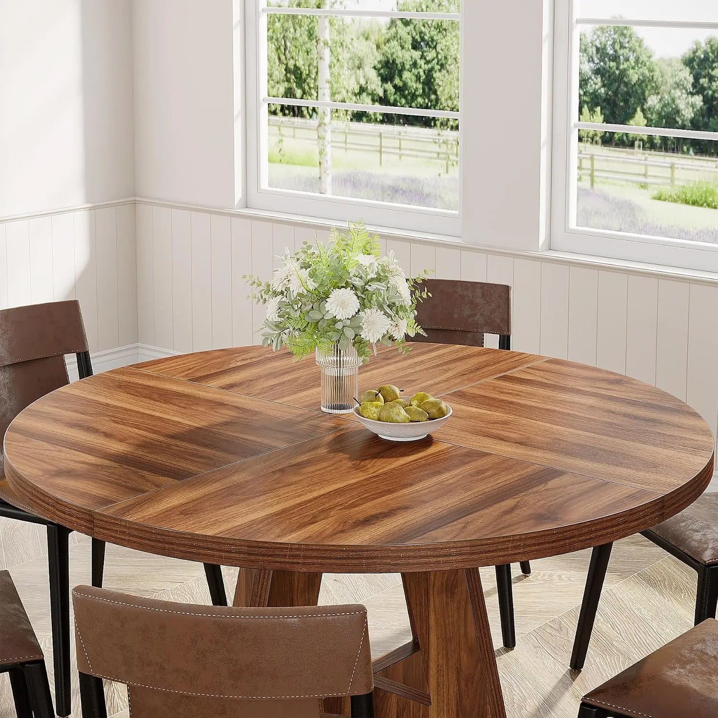 Round wooden dining table with chairs in a bright room with large windows.