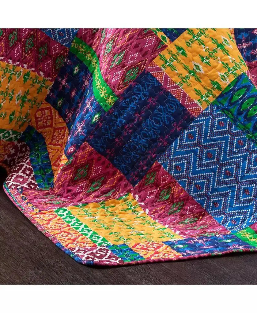 A close-up of a colorful patchwork quilt draped over a dark wooden floor, featuring vibrant blocks of pink, blue, yellow, green, and orange with various geometric and floral patterns.