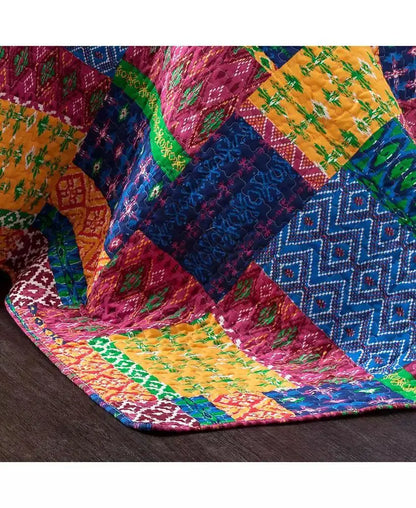 A close-up of a colorful patchwork quilt draped over a dark wooden floor, featuring vibrant blocks of pink, blue, yellow, green, and orange with various geometric and floral patterns.