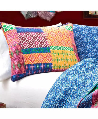 A colorful patchwork quilted pillowcase with vibrant geometric and ikat patterns in pink, orange, yellow, green, and blue, styled on a bed alongside a matching blue patterned quilt.