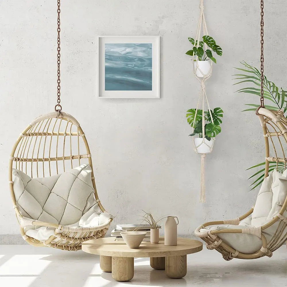 Two hanging rattan chairs with cushions in a room with a table, plants, and a framed picture on the wall.