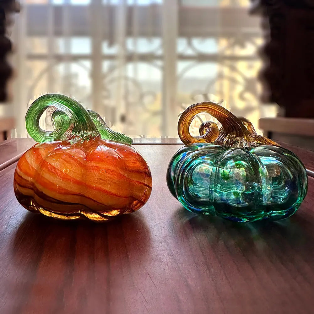 Two glass pumpkins, one orange and one green, on a wooden surface with a blurred background.