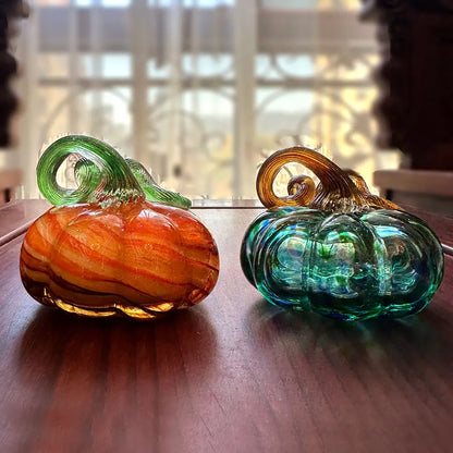 Two glass pumpkins, one orange and one green, on a wooden surface with a blurred background.
