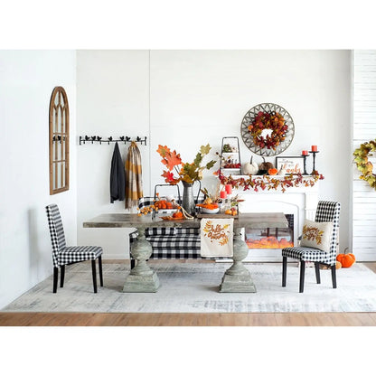 A styled dining room scene with a rustic wooden table, black-and-white checkered chairs, a white rug, and fall decor including pumpkins, autumn leaves, a wreath, and a mantel display against white walls.