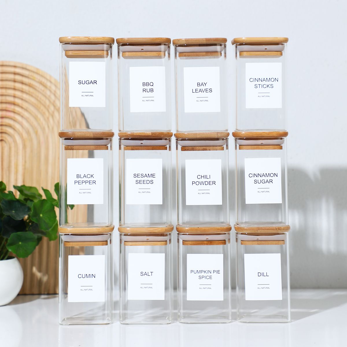 Set of clear spice jars with wooden lids on a white surface, featuring labels for various spices.