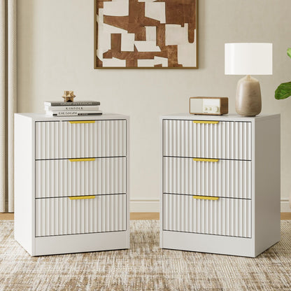 Two white three-drawer nightstands with vertical fluted drawer fronts and slim gold handles, shown in a styled room setting with books, a lamp, a radio, and wall art.