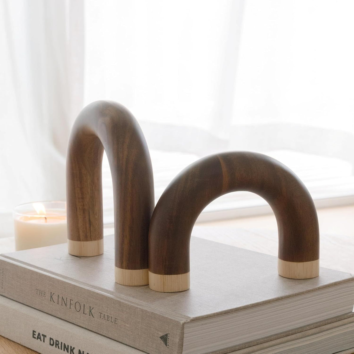 Two curved dark wood bookends with light wood ends, displayed on top of stacked books in a softly lit room with a candle and window in the background.