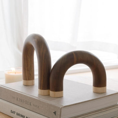 Two curved dark wood bookends with light wood ends, displayed on top of stacked books in a softly lit room with a candle and window in the background.