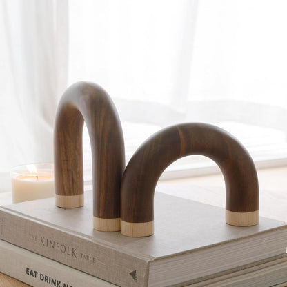 Two curved dark wood bookends with light wood ends, displayed on top of stacked books in a softly lit room with a candle and window in the background.