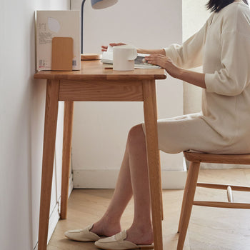 Person sitting at a wooden desk with a cup, laptop, and books in a minimalistic room.